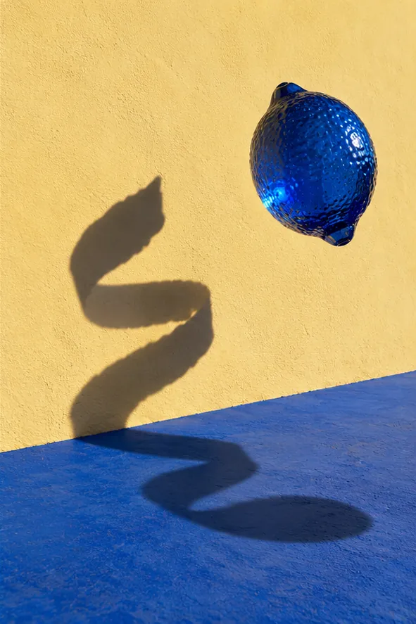 A blue glass lemon floats in front of a yellow wall, while its shadow wanders off in a long, wavy line.