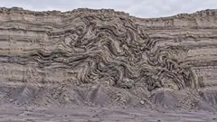 A grey badlands cliff is neatly layered at the top and bottom, and in the middle the strata twist and loop into a knot. A few million years went sideways.