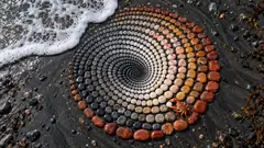 On a black sand beach, pebbles are laid in a perfect spiral graded from dark to cream to red, and a small orange crab sits at the edge, adjusting one. The sea is coming for it anyway.