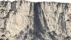 A tall pale limestone cliff has a dark, waterfall-shaped shadow running down its face and spreading at the base like spray, with no water anywhere. The waterfall left and its shadow stayed.