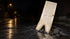 On a wet street at night, a white panelled door pushes up through the cracked asphalt on two roots, like a tooth coming in. The road will be tender for a while.